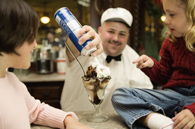 A man is squirting whipped cream from a blue can onto a hot fudge sundae as a child and woman look on